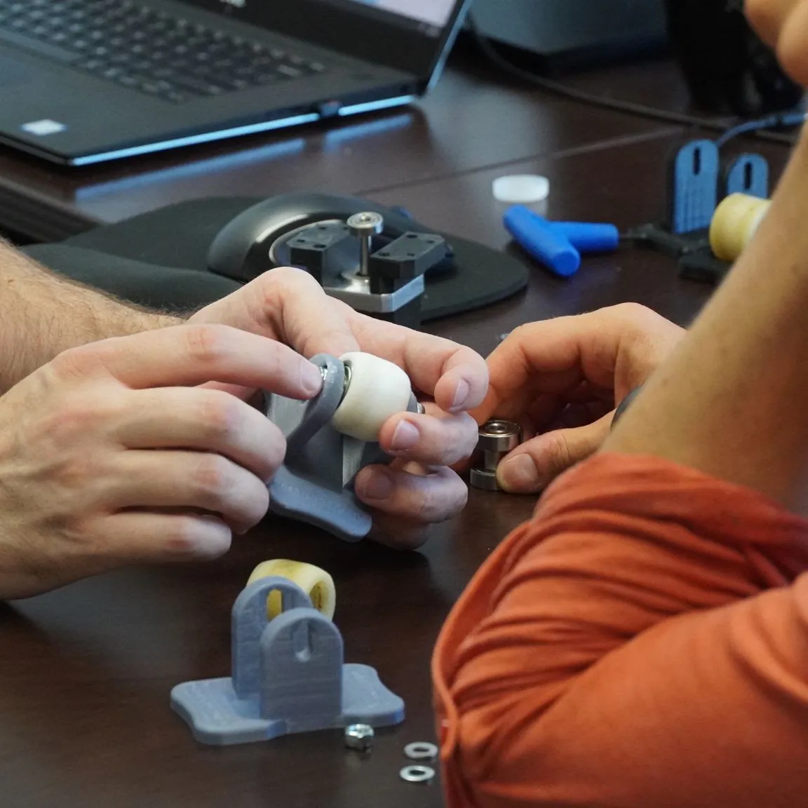 Hands assembling blue 3D-printed prototype parts on a shop bench, with more printed pieces stacked nearby.