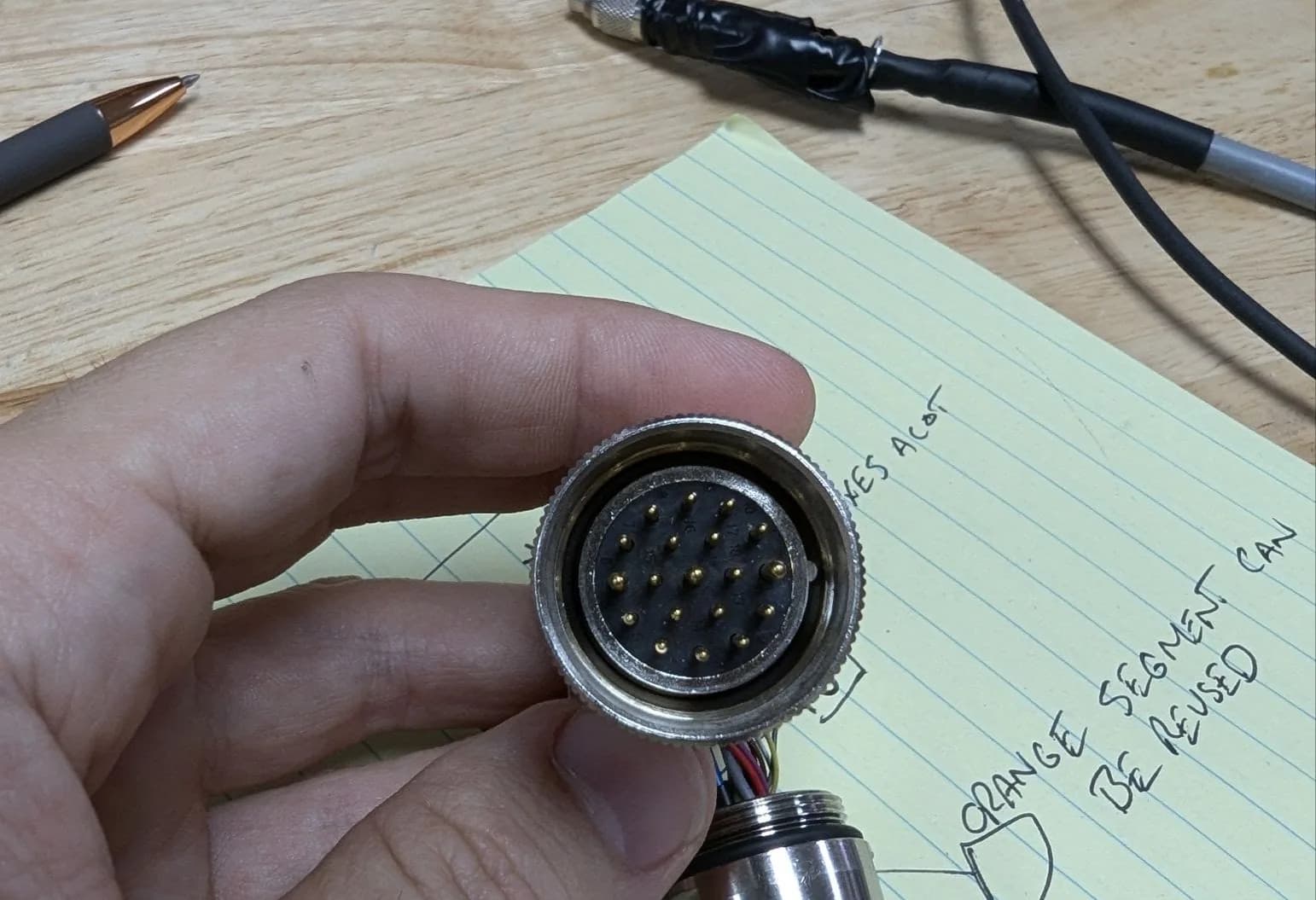 A hand holding a circular multi-pin connector, with a pinout sketch on the bench behind it.