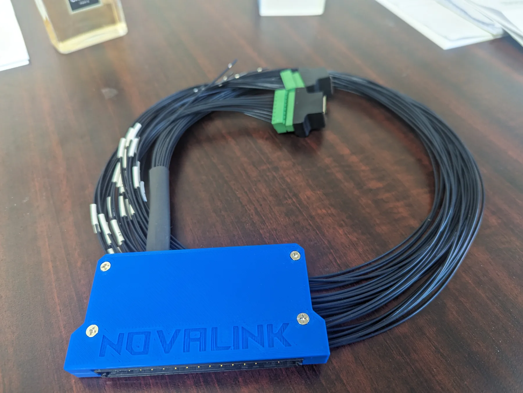 A blue 3D-printed connector housing, with NOVALINK molded into the cover, on a coiled black harness with green terminal connectors.