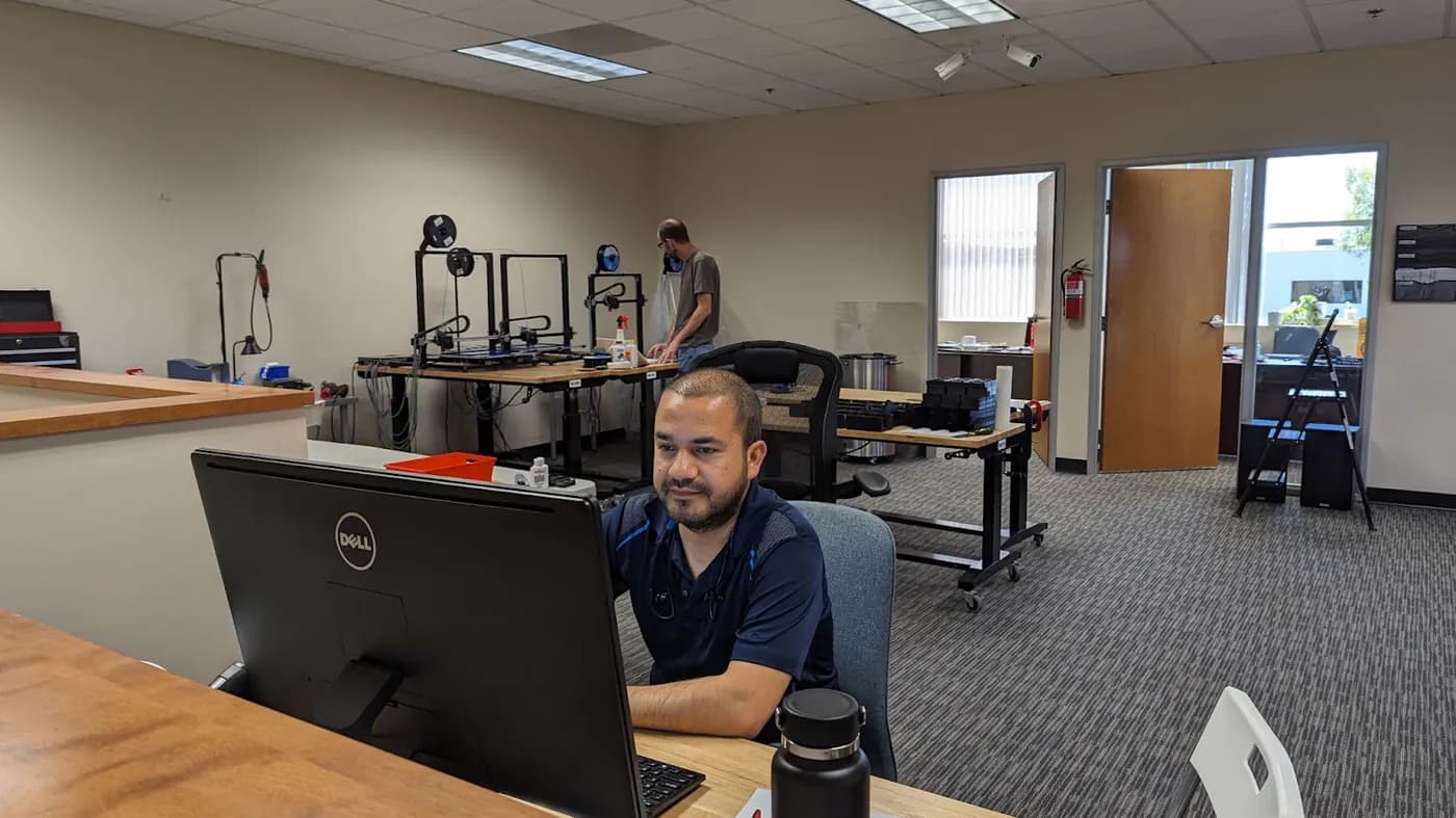 An engineer working at a computer in the shop, with a rack of desktop 3D printers behind him.