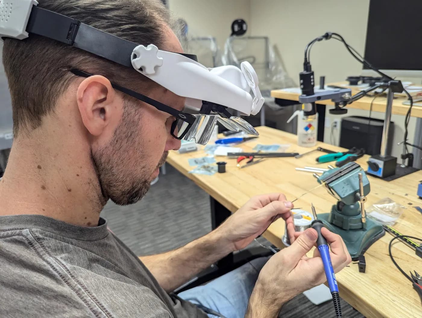 An engineer at a white bench soldering a wired assembly under a task lamp, with power supplies and hand tools beside the work.