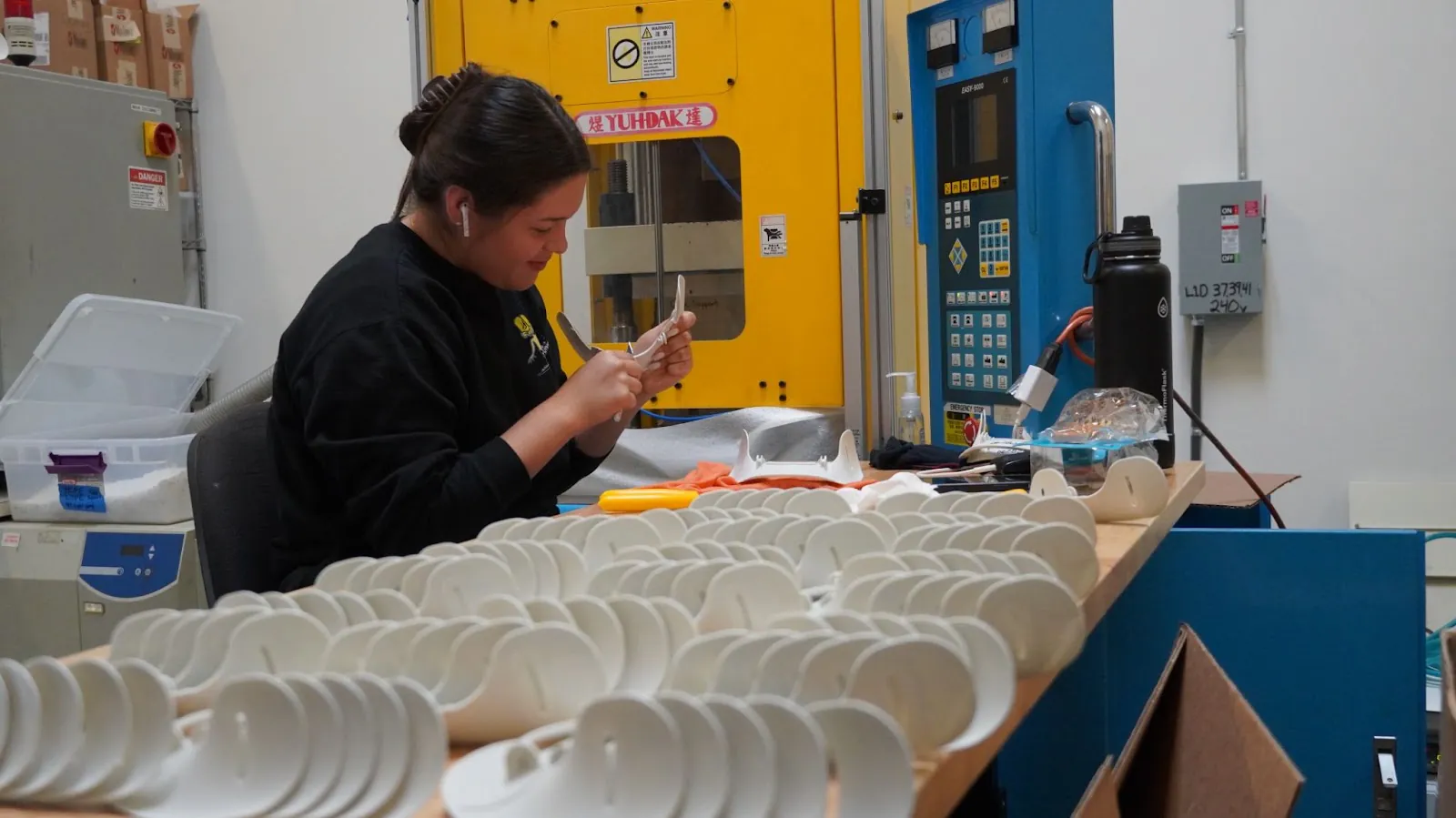 An inspector at a vertical molding machine checking a small molded part, with finished housings on the press table.