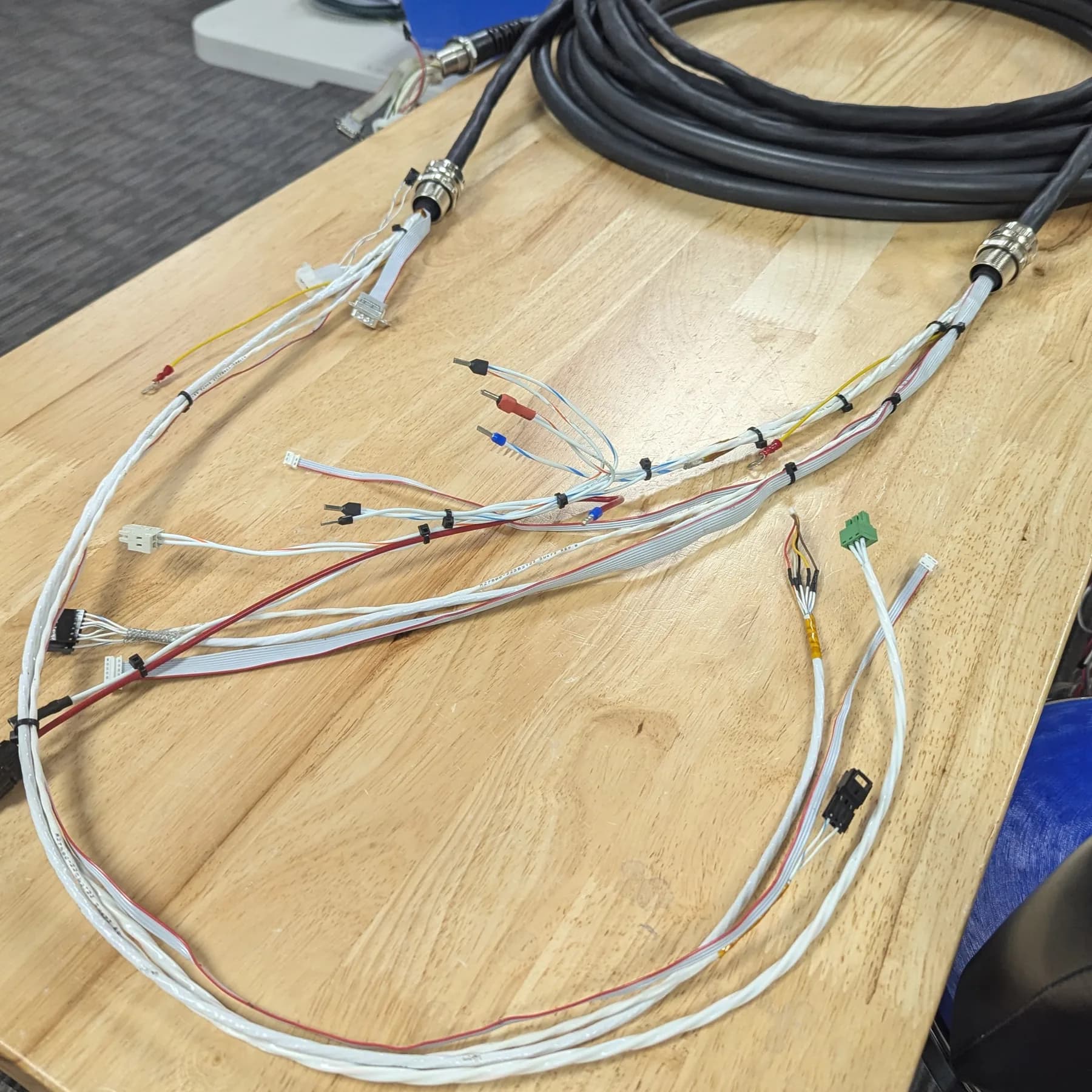 A multi-branch wire harness on a wood bench, with circular metal backshells, a ribbon breakout, and crimped and ferrule terminations.