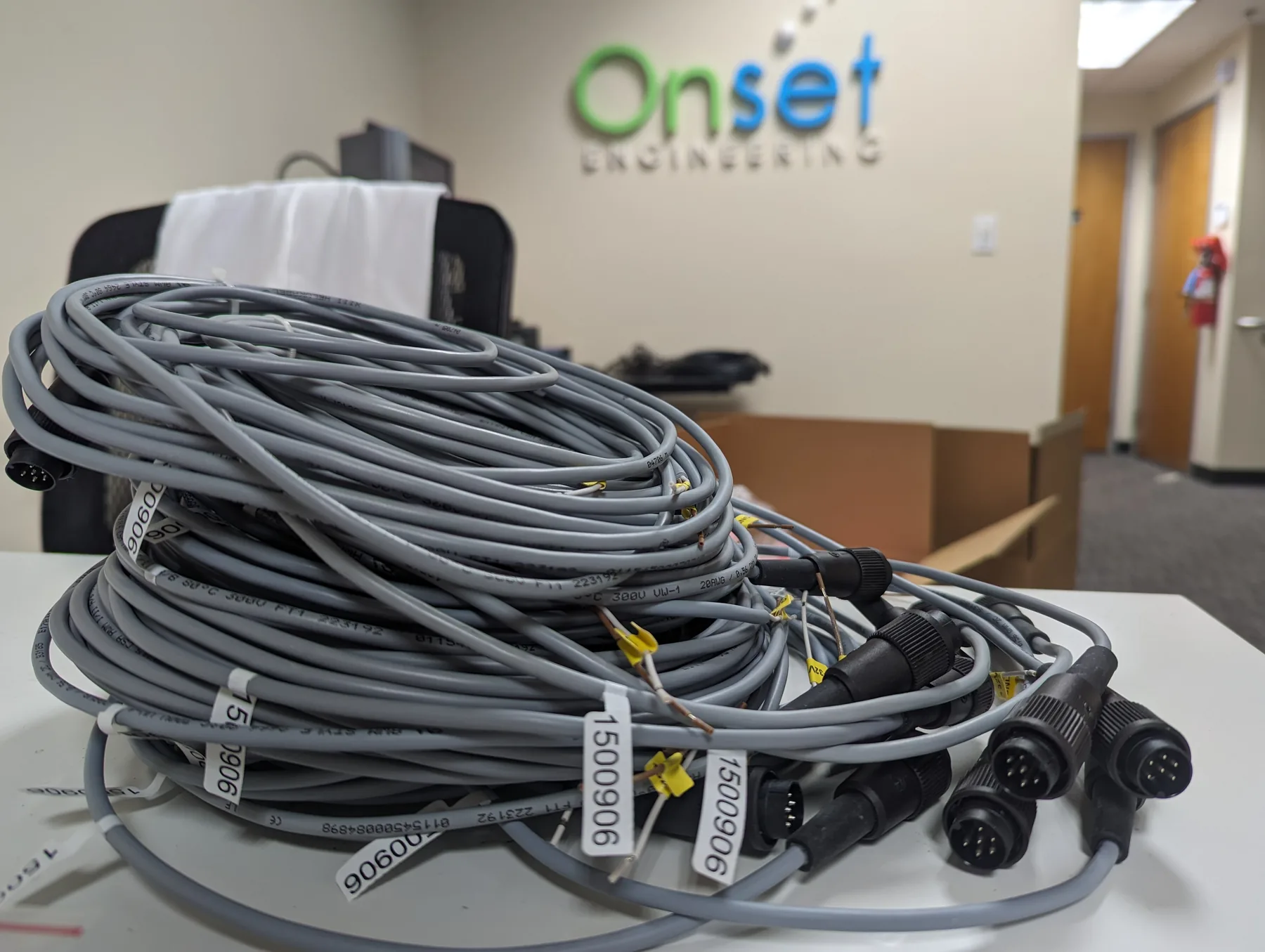 A coil of gray overmolded multi-pin circular cables with part labels, on a table in front of the Onset Engineering wall sign.