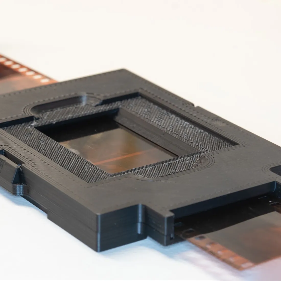 A 3D-printed film-holder fixture with a blue geared roller, a gray frame, and two mounting brackets.
