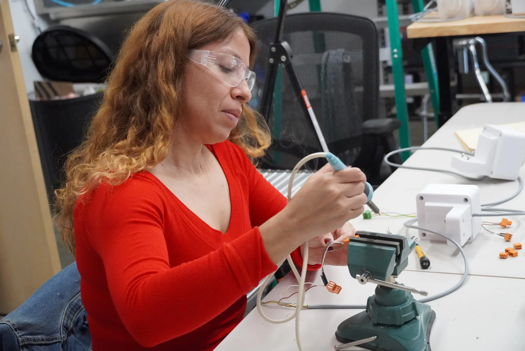 A technician in safety glasses soldering a wire assembly held in a bench vise, with white enclosures on the table.